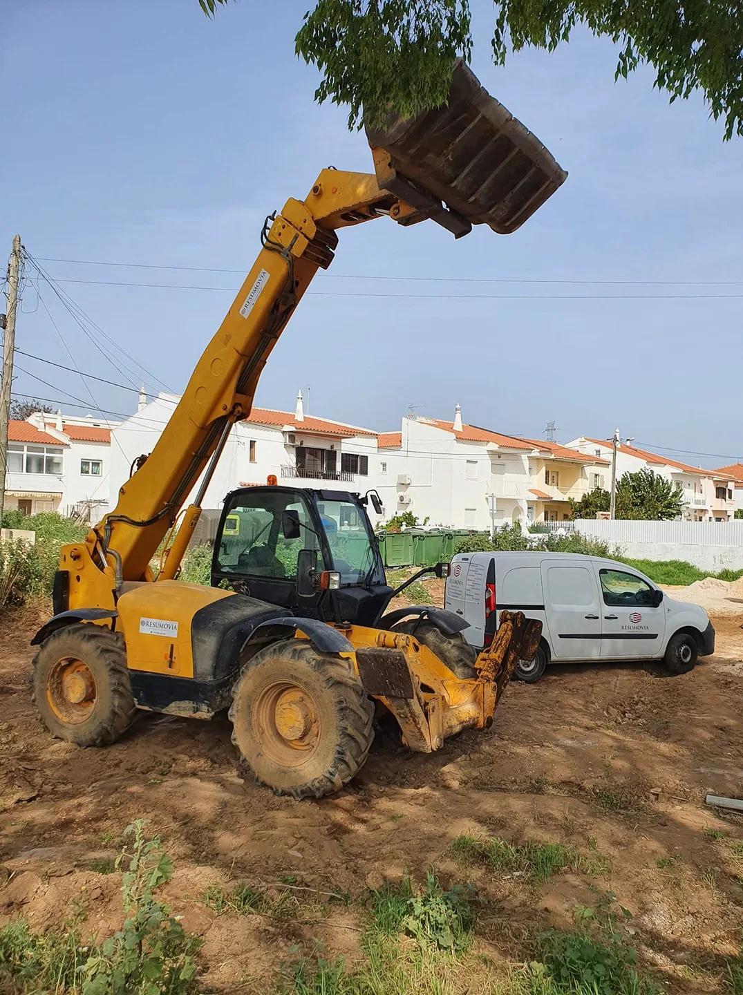 Empilhador telescópico a carregar num terreno, com moradias ao fundo